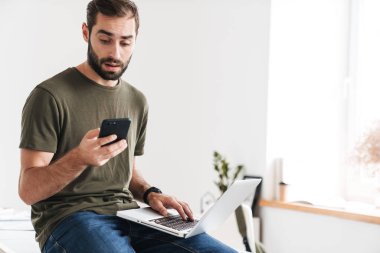 Yakışıklı, şaşırmış bir adamın cep telefonuyla yazı yazması ve dizüstü bilgisayar kullanması