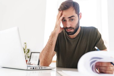 Laptop kullanan ve doküman okuyan kafası karışmış genç bir adamın resmi
