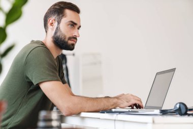 Masada otururken dizüstü bilgisayarda yazı yazan beyaz bir genç resmi