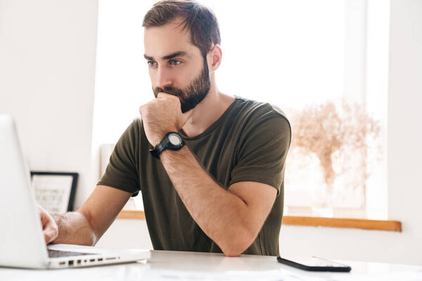 Image of handsome thinking man typing on laptop while sitting