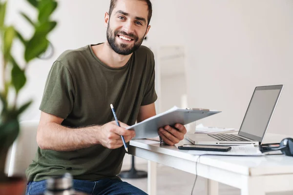 Bild eines lächelnden jungen Mannes mit Laptop und Klemmbrett — Stockfoto