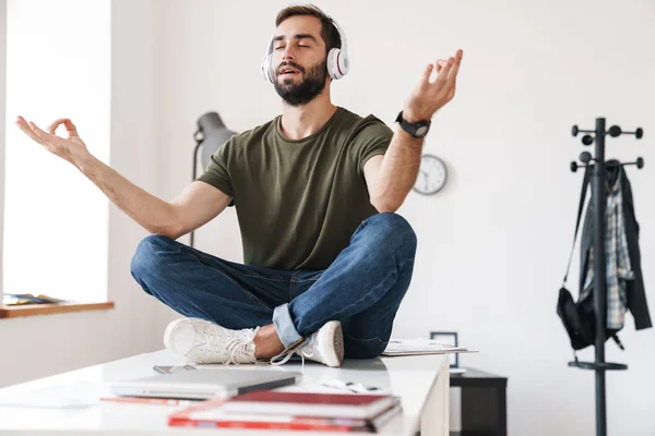 Imagen del hombre tranquilo escuchando música con auriculares mientras está sentado en el escritorio —  Fotos de Stock