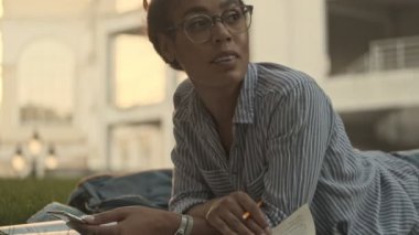 Gözlüklü düşünceli, güzel Afrikalı bir kadın akıllı telefon tutuyor ve parktaki ekosenin üzerinde yatarken gözlerini kaçırıyor.