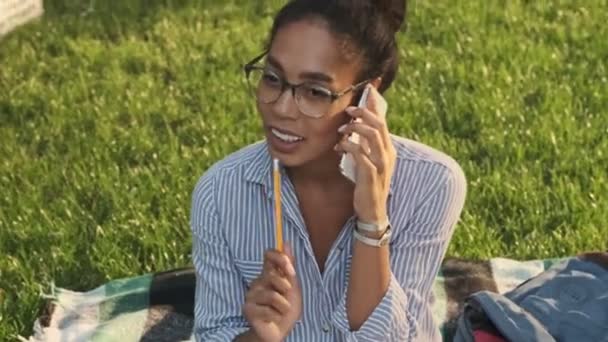 公園に座っている間に手に鉛筆でスマートフォンで話している眼鏡で幸せなアフリカの女性 — ストック動画