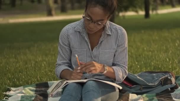 公園に座っている間に雑誌や何かを書くことで勉強している眼鏡の中で幸せなアフリカの女性 — ストック動画