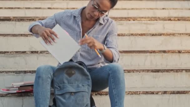 Спокійна Африканська Жінка Окулярах Кладе Блокнот Рюкзак Сидячи Сходах Відкритому — стокове відео