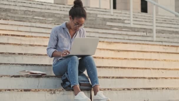 屋外の階段に座っている間 ラップトップコンピュータを使用して眼鏡できれいなアフリカの女性 — ストック動画