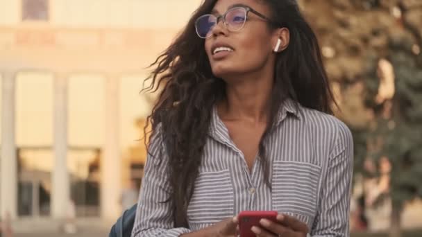 通りに立ちながら 遠くを見てスマートフォンを使っている眼鏡やイヤフォンを手にしたアフリカ人の女性が — ストック動画