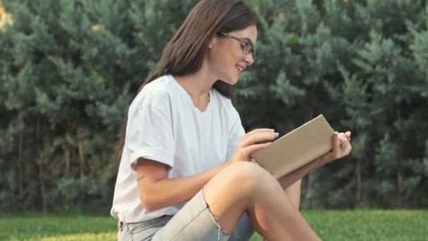 Όμορφη Νεαρή Κοπέλα Φορώντας Γυαλιά Διαβάζει Ένα Βιβλίο Ενώ Κάθεται — Αρχείο Βίντεο