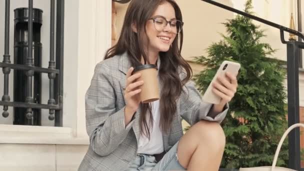 在市中心 一个戴着眼镜 面带微笑的女孩坐在那里 一边喝着咖啡 一边用智能手机 — 图库视频影像