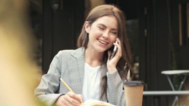 Şirin, gülen genç bir kadın dışarıda kafede telefonla konuşurken defterine bir şeyler mi yazıyor? Açık havada.