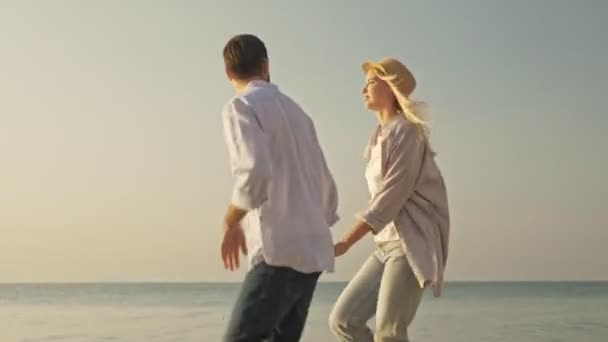 明るい笑顔のカップルの男と女が走っていて 夜明けにビーチで回転している — ストック動画