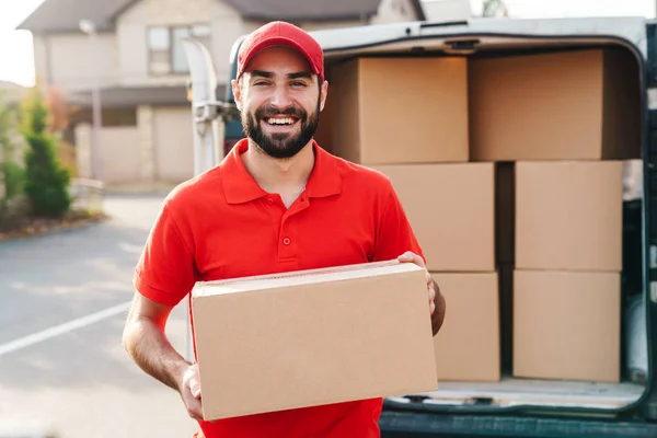 Imagen del joven repartidor sonriente de pie con caja de paquetes — Foto de Stock