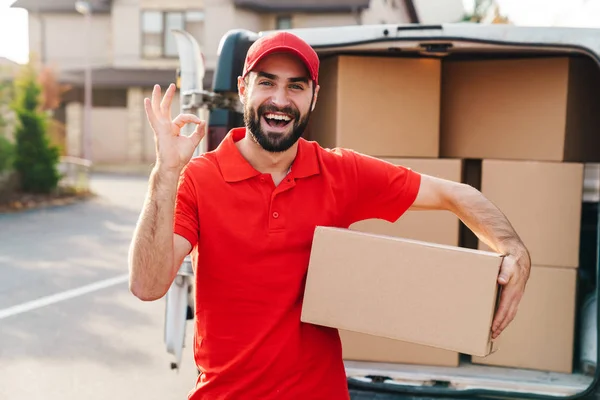 Imagem de um homem de entrega de pé com caixa de encomendas e mostrando sinal ok — Fotografia de Stock