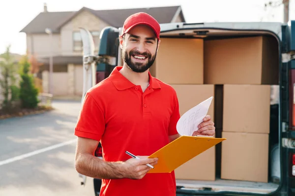 Image of cheerful delivery man holding clipboard and writing — 스톡 사진