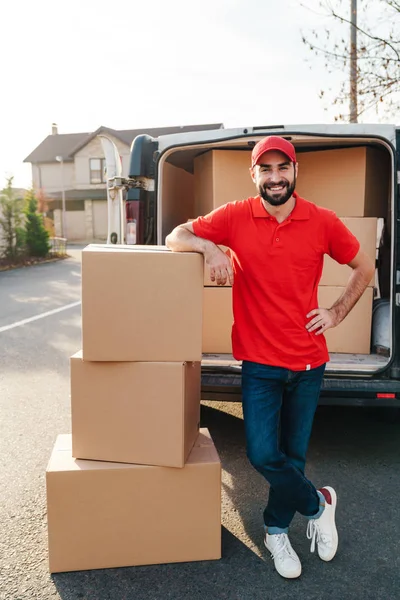 Image of smiling young delivery man standing with parcel boxes near car — Stock Photo, Image