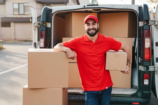 Bild av glad ung leverans man stående med paket lådor nära bilen — Stockfoto