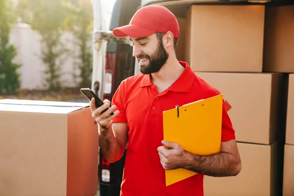 Image of happy delivery man holding clipboard and using cellphone