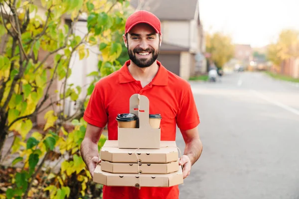 Imagen del joven repartidor sonriente sosteniendo cajas de pizza y café — Foto de Stock