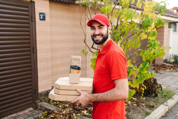 Image of cheerful young delivery man holding pizza boxes and coffee