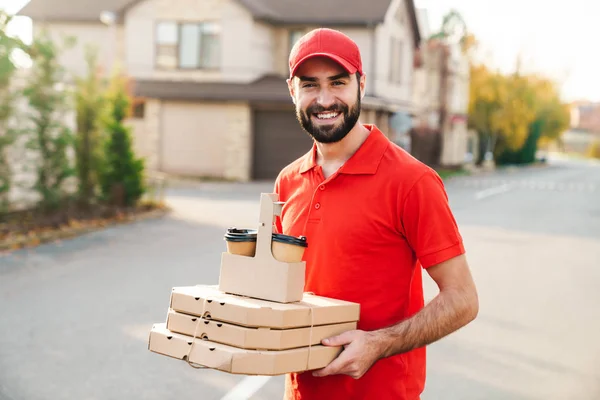 Imagen del joven y alegre repartidor sosteniendo cajas de pizza y café — Foto de Stock