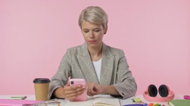 Şık bir kadın akıllı telefonundan bir şey izliyor ve sonra pembe arka planda masanın başında otururken kendisini mutlu eden iyi haberi alıyor.