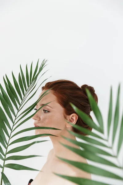 Genç kızıl saçlı kadın yaprak yeşil çiçekleri ile beyaz duvar arka plan üzerinde izole poz. — Stok fotoğraf