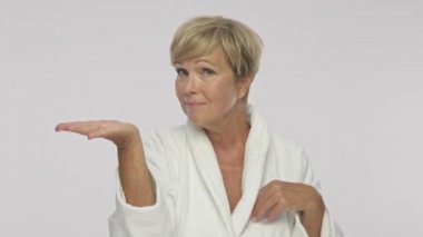 A pleased adult woman with short blond hair wearing white housecoat holding copyspace on her palm isolated over white background