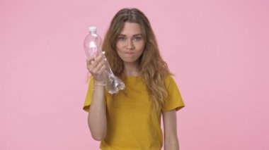 Irritated caucasian woman in yellow t-shirt is holding plastic bottles while crumpling them isolated over pink background