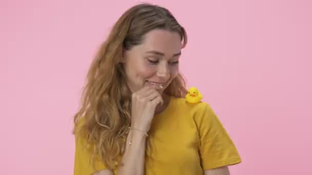 一位年轻貌美的女士正在玩一个小黄鸭玩具 玩具放在肩上 与粉红的背景隔离开来 — 图库视频影像