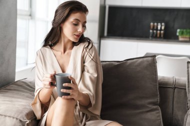 Evdeki kanepede oturup çay içen genç bir kadın resmi.