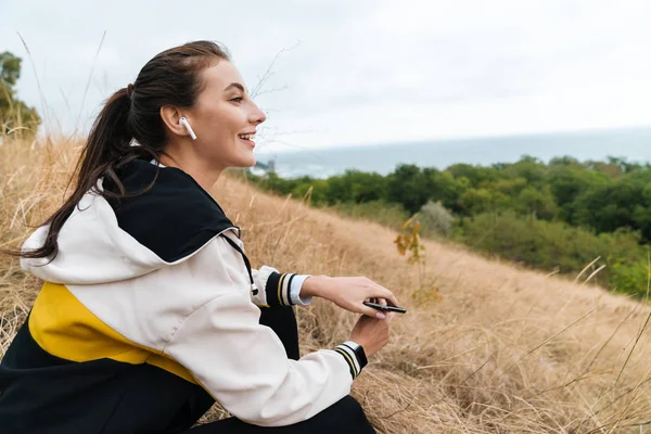 Zdjęcie miłej szczęśliwej kobiety słuchawki i smartfon podczas siedzenia — Zdjęcie stockowe