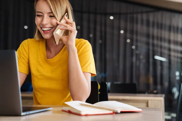 Foto einer fröhlichen jungen Frau, die Laptop benutzt und mit dem Handy spricht — Stockfoto