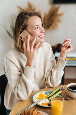 Kahvaltı yaparken cep telefonuyla konuşan memnun bir kadının fotoğrafı.