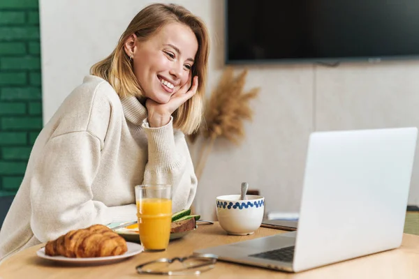 Foto de mujer rubia alegre usando el ordenador portátil mientras desayunaba — Foto de Stock