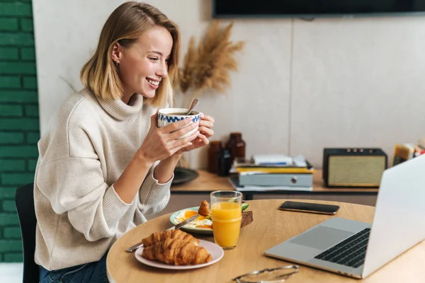 Foto de la mujer rubia feliz usando el ordenador portátil mientras desayuna — Foto de Stock