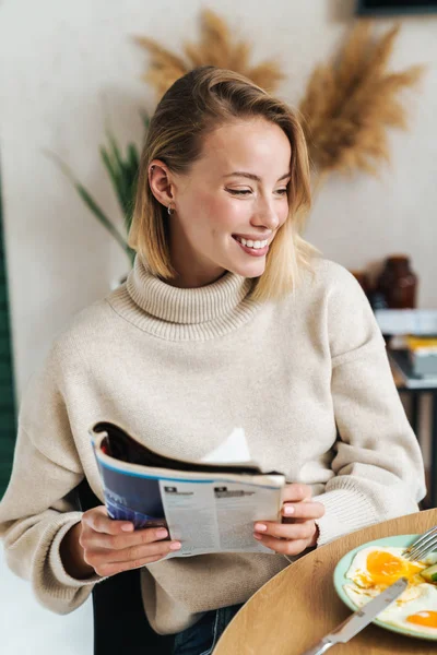 Φωτογραφία από χαρούμενη ξανθιά γυναίκα ανάγνωση περιοδικό, ενώ έχοντας πρωινό — Φωτογραφία Αρχείου