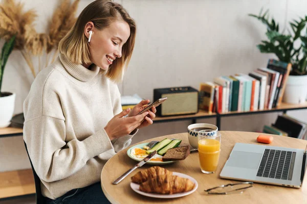 Fénykép egy nőről, aki mobilt és fülhallgatót használ reggeli közben — Stock Fotó