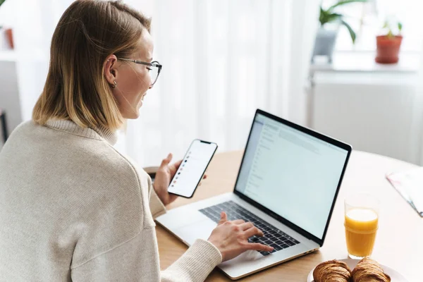 Photo of happy caucasian woman typing on laptop and cellphone — 스톡 사진