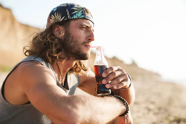 Foto di pensare giovane bevendo soda e seduto sulla spiaggia soleggiata — Foto Stock