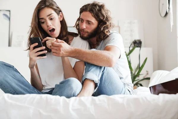 Couple indoors at home sit on bed using mobile phone.