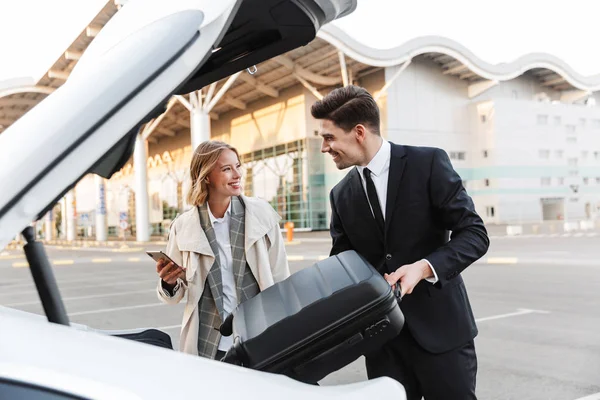 Image of young businesslike man and woman putting luggage in car — ストック写真