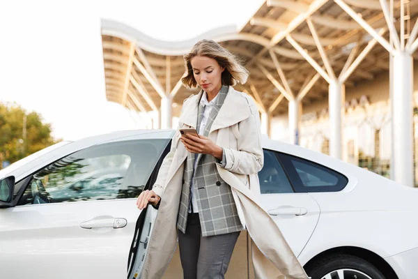 Foto de una joven mujer de negocios usando el teléfono celular mientras sale del coche —  Fotos de Stock