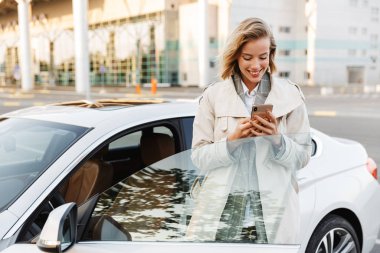 Image of businesslike woman standing by car and using cellphone 