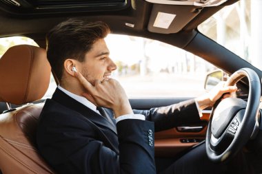 Image of young businesslike man in suit using earbuds while driv