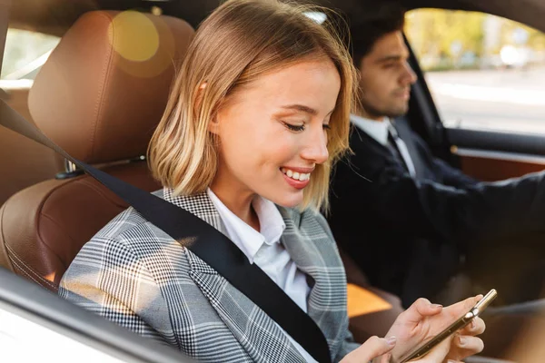 Foto del hombre y la mujer usando el teléfono celular y sonriendo mientras conduce en coche — Foto de Stock