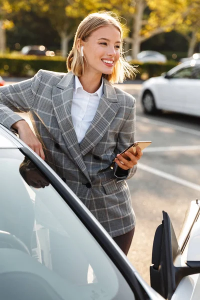 Foto von Geschäftsfrau mit Kopfhörer und Handy, während sie sich auf Auto lehnt — Stockfoto