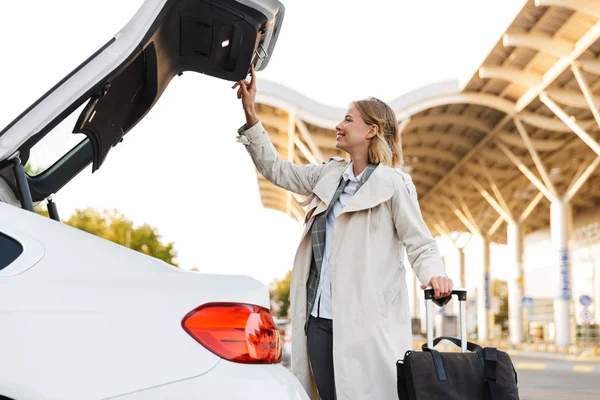 Photo of smiling businesswoman holding suitcase and opening car trunk — Stockfoto