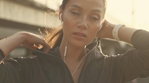 閉鎖ビューの静かなアジアのスポーツ女性でイヤフォンにフードを置くと離れて屋外を見ます — ストック動画