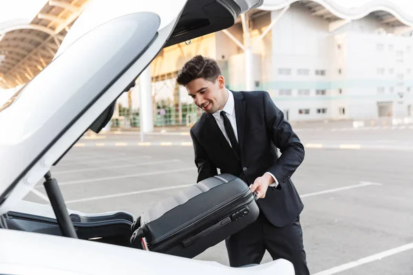 Photo of smiling businessman putting suitcase in car trunk